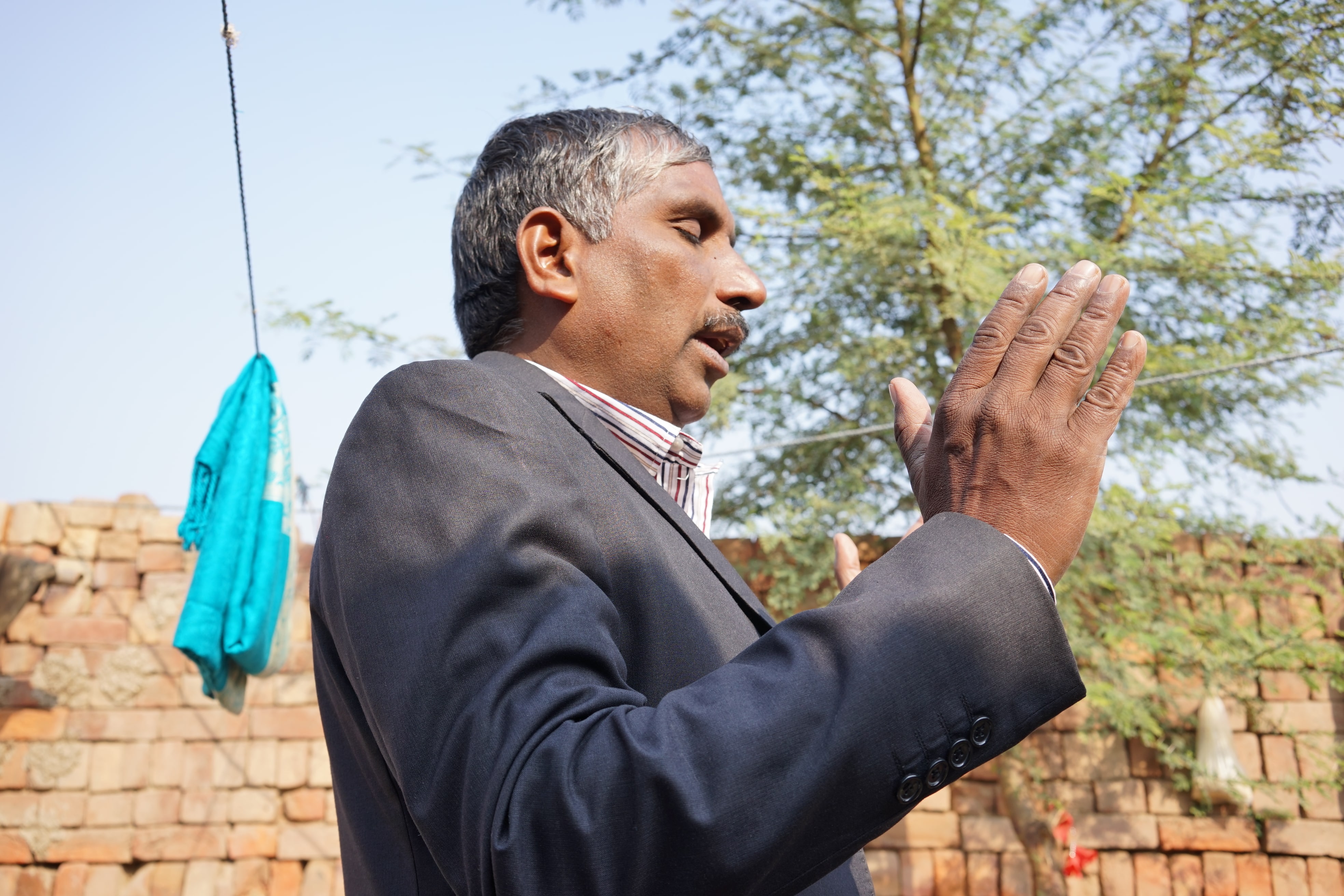 A man in a suit gestures with his hands while speaking outdoors, with a brick wall and tree in the background.