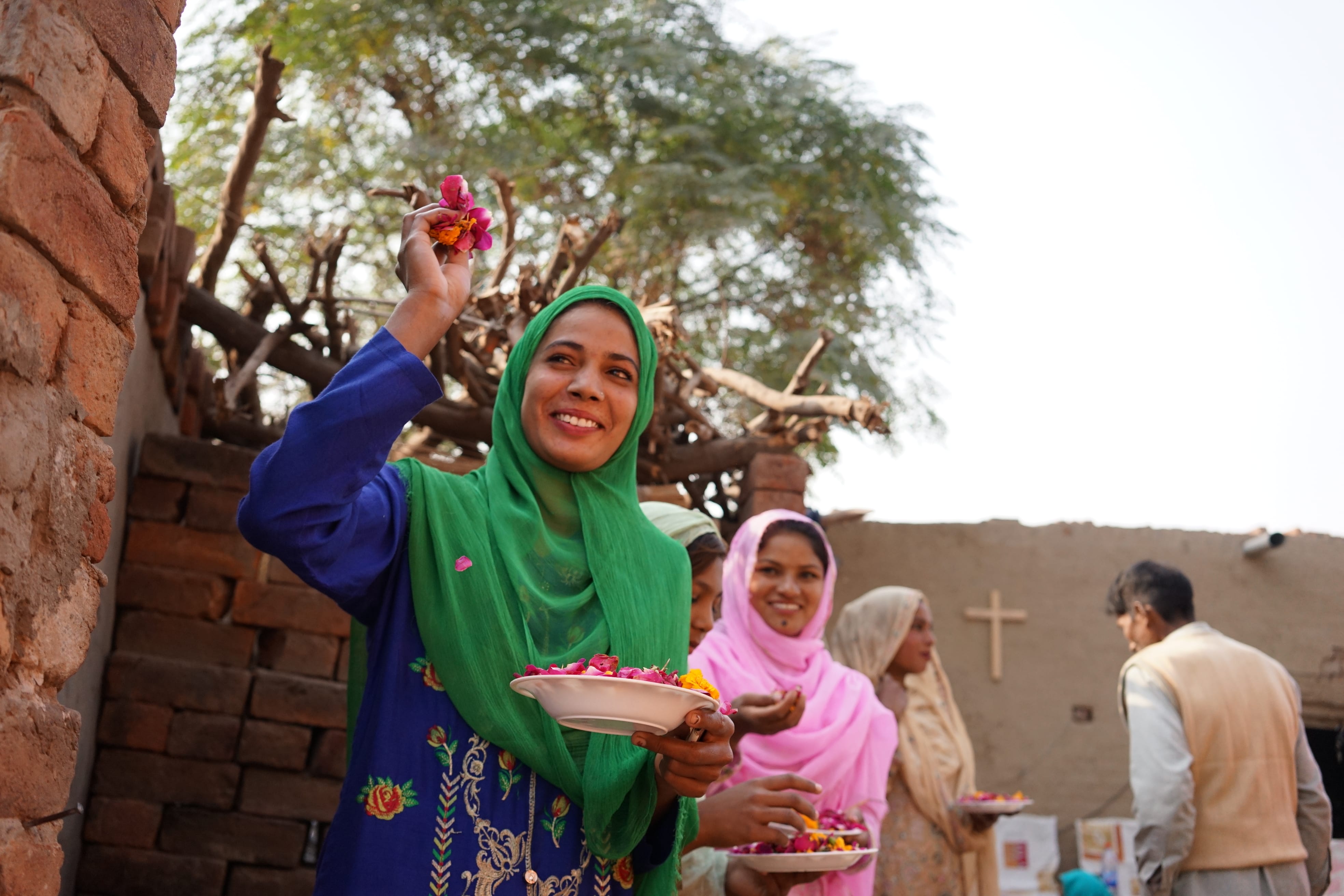 A smiling woman in a green headscarf holds a plate of flowers, with others around her, in an outdoor setting near a brick wall.