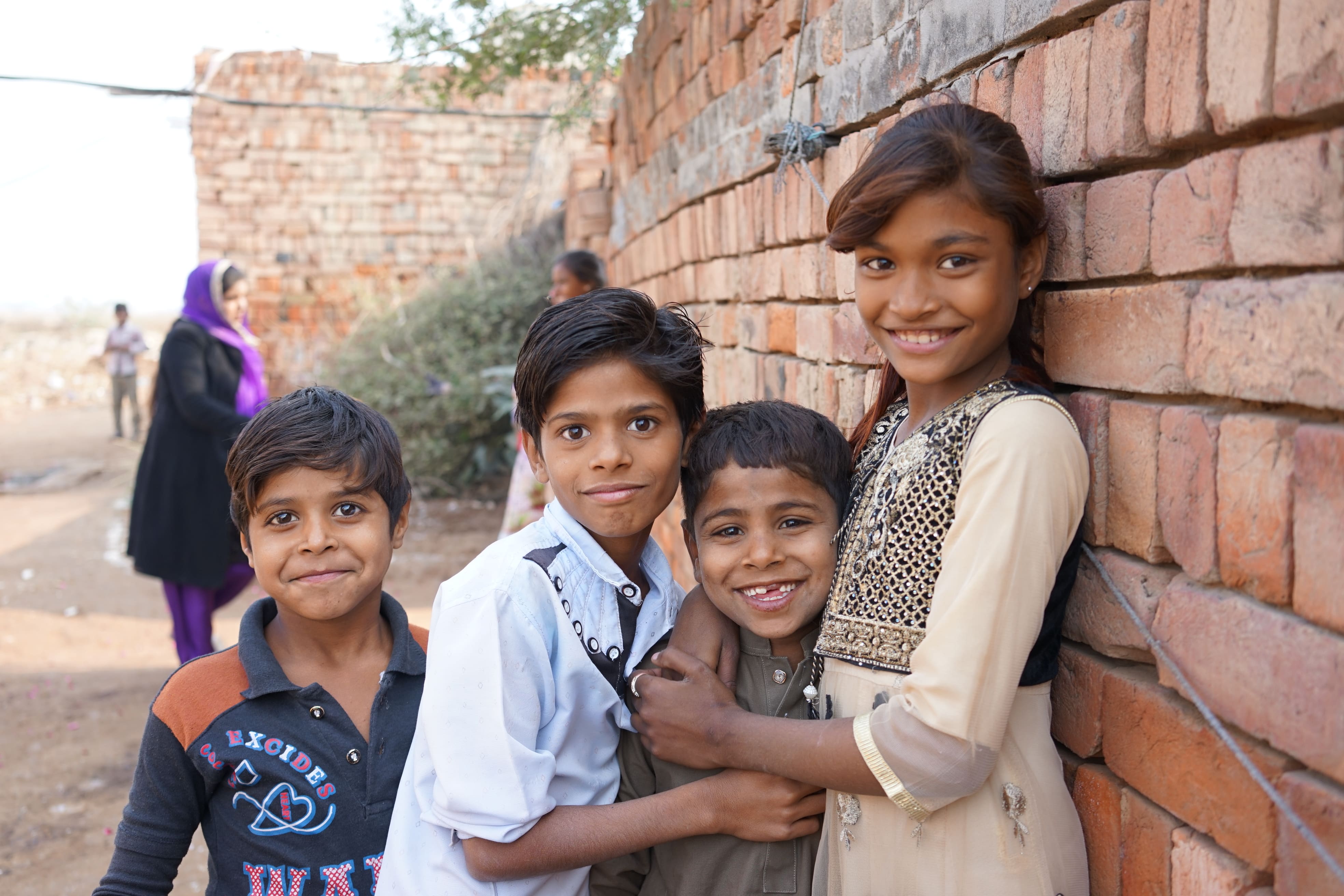 Four smiling children stand close together against a brick wall, with a few people and more bricks in the background.