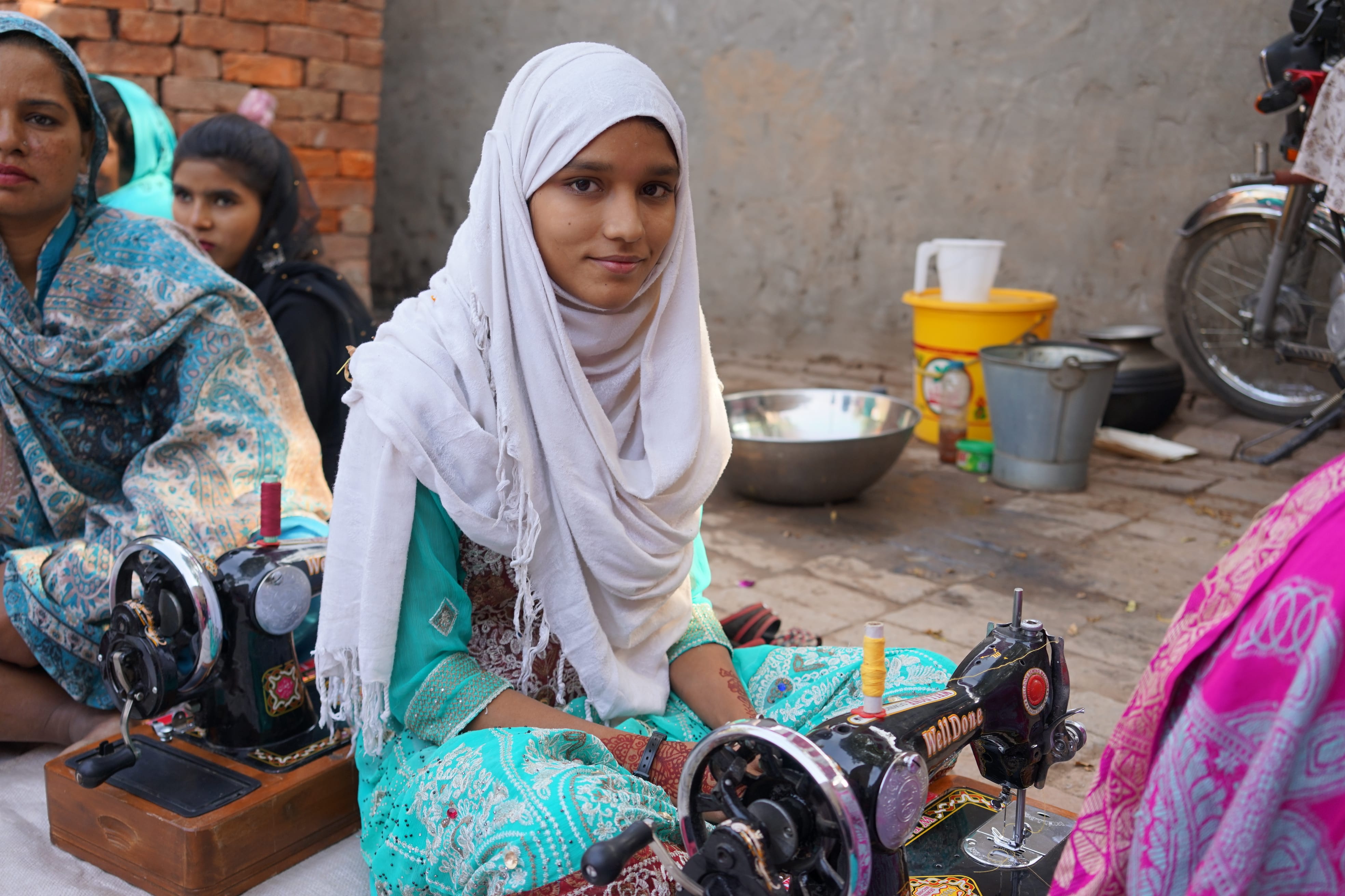 A young girl in a white headscarf and turquoise outfit sits at a sewing machine, with other women around her. 