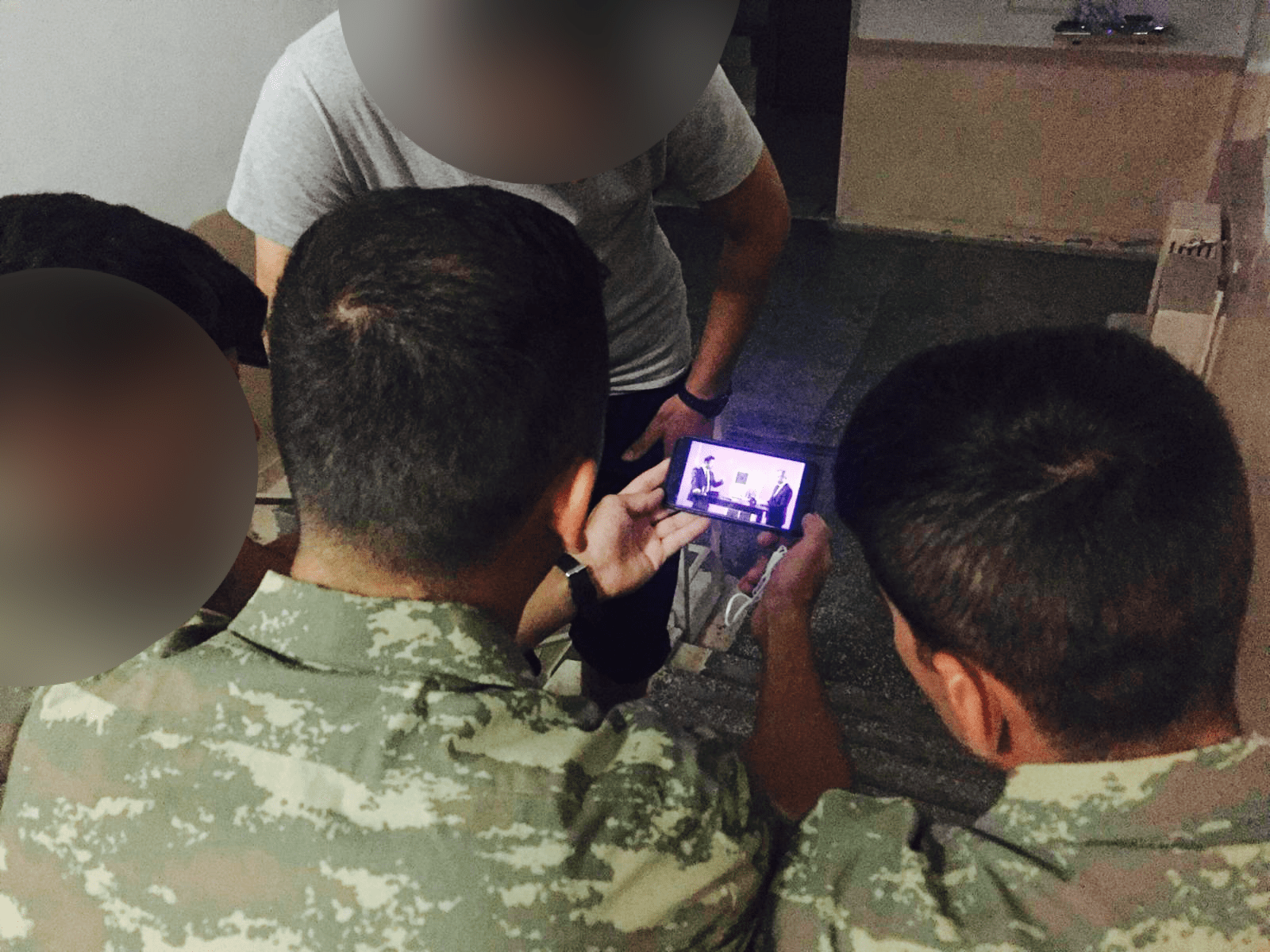 A group of army individuals huddled around a phone watching an interview. 