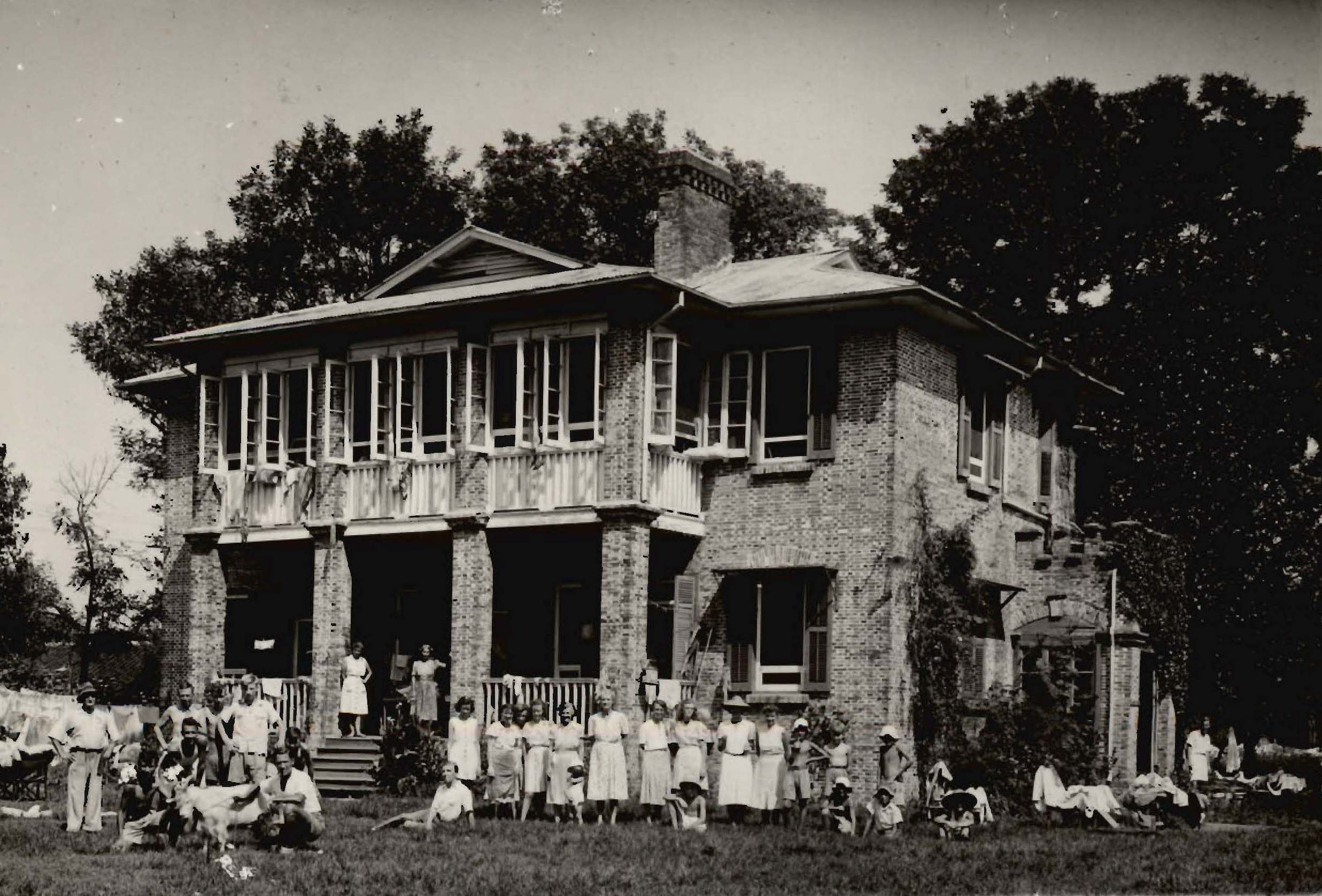 A vintage photograph of a large brick house with many people, including children, gathered in front of it, set in a lush, tree-filled landscape.