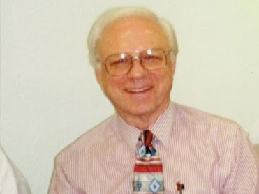 An elderly man with white hair and glasses is smiling at the camera. He is wearing a light pink, striped dress shirt and a colourful, patterned tie. The background is plain and light-coloured.