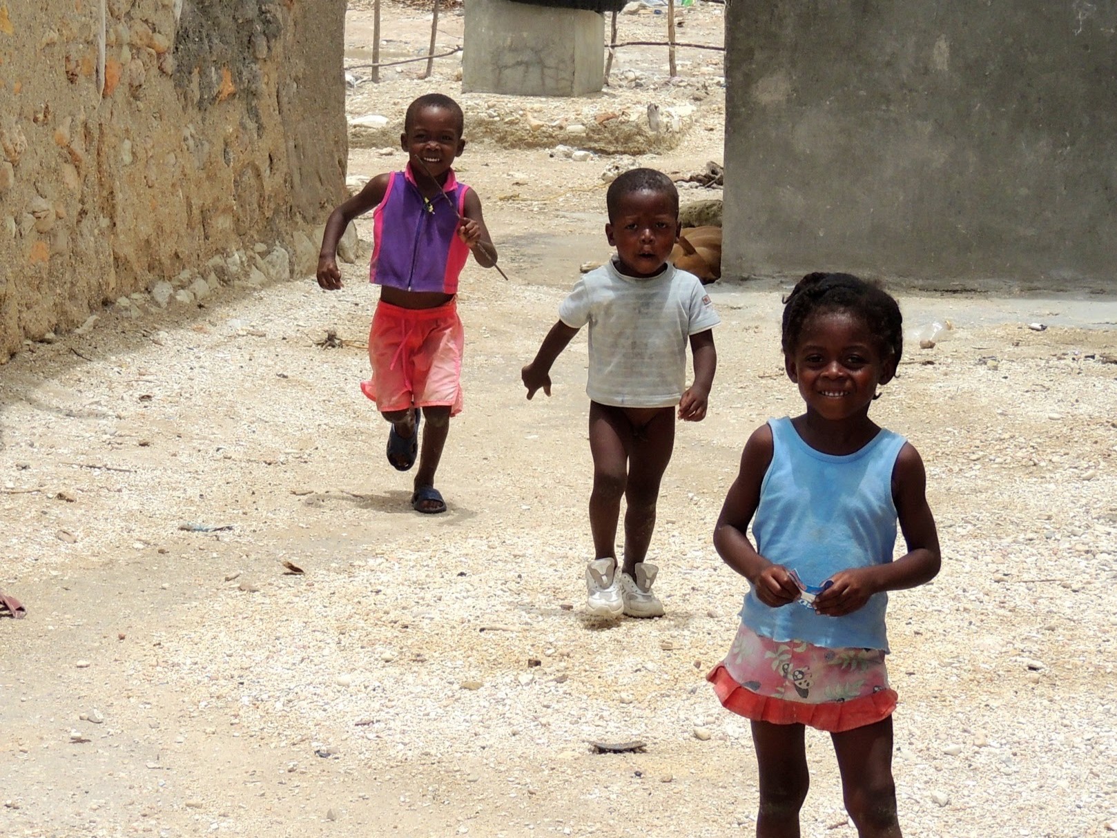 Three young children are playing and running on a dirt path between stone and concrete buildings. 