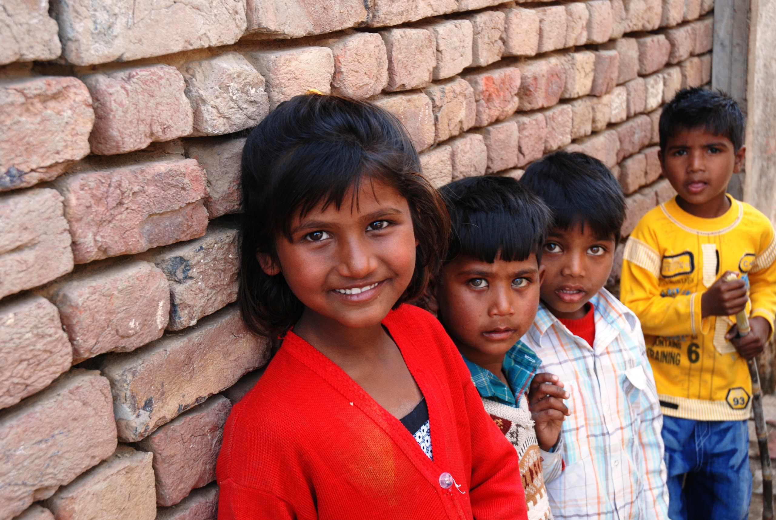 Four young children are standing against a brick wall, smiling and looking at the camera.