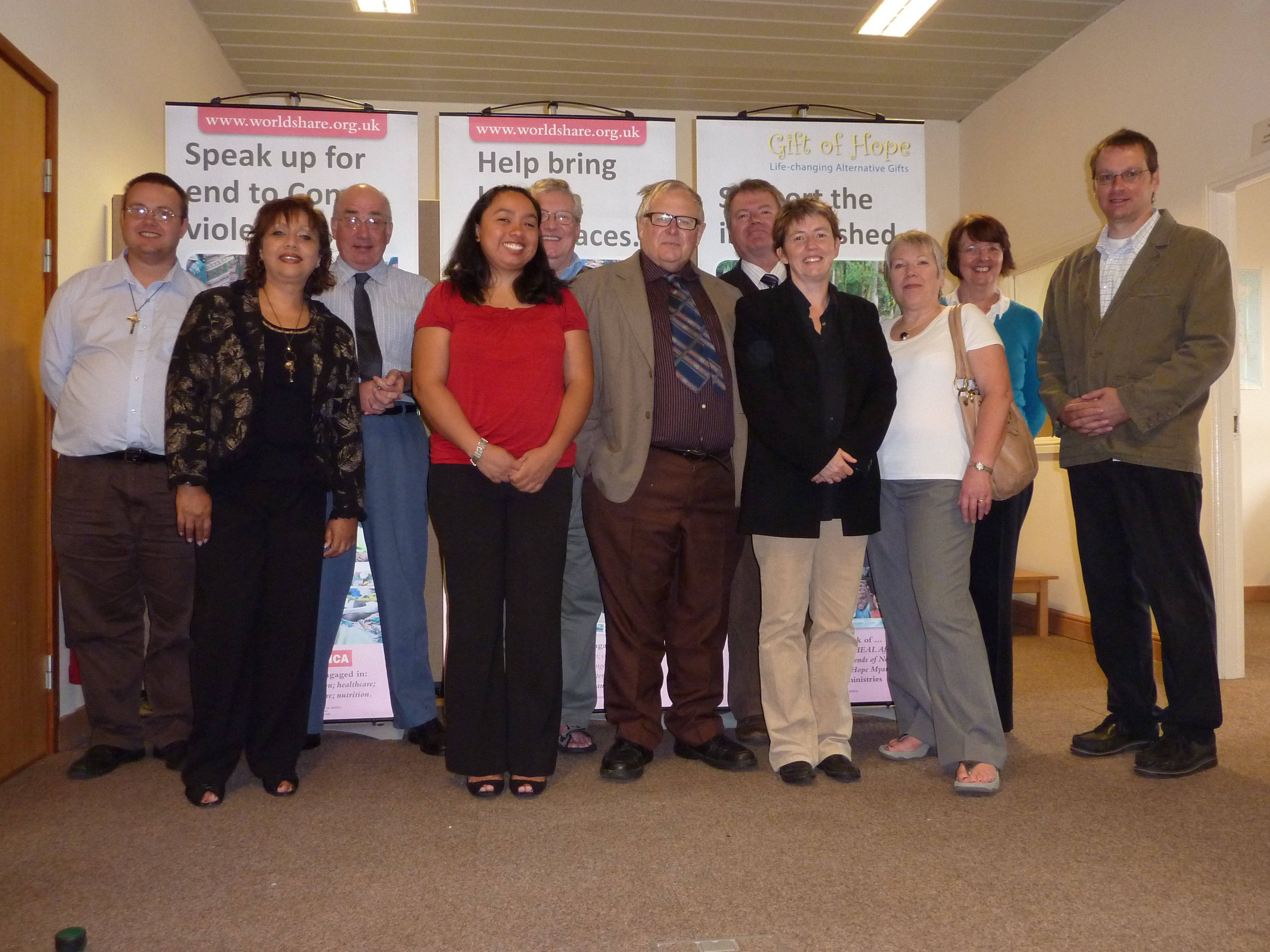 A group of ten adults is standing together indoors, posing for a photo in front of banners from worldshare.org.uk. 