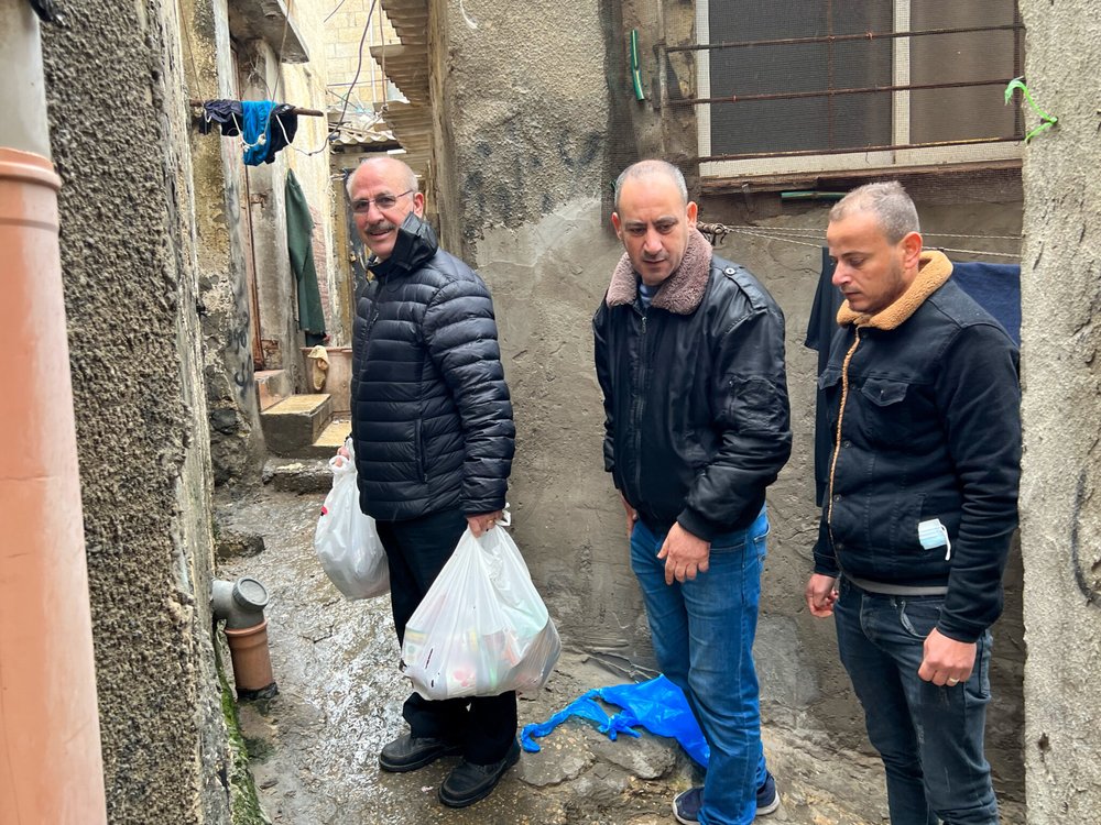 Three men stand in a narrow alleyway holding bags of supplies, wearing jackets on a chilly day. 