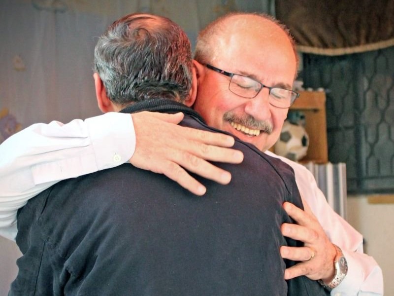 Two men warmly embrace, smiling with their eyes closed, in a heartfelt moment indoors.