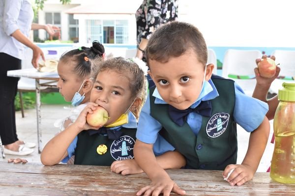 Two young children in school uniforms sit at a table, with one girl eating an apple and a boy looking curiously at the camera