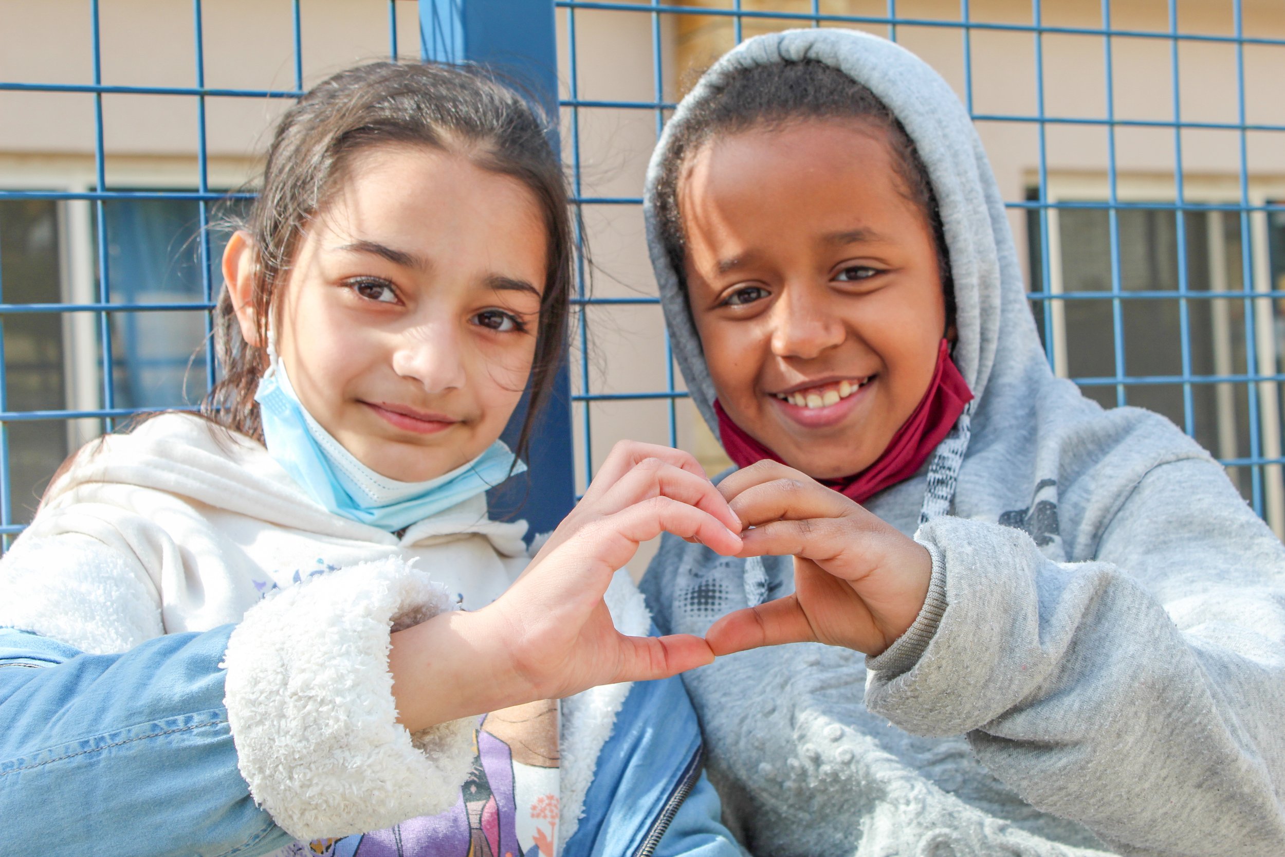 Two young girls smile and form a heart shape with their hands, wearing casual jackets and standing in front of a blue metal fence.