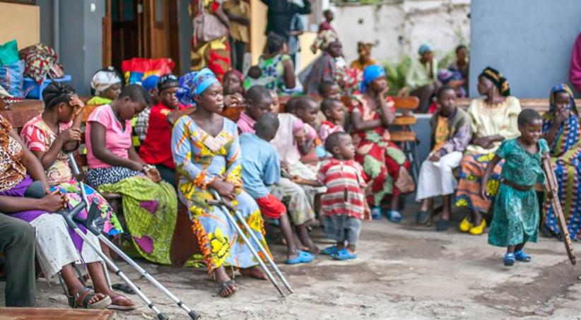 a large group of people sitting outside, a mixture of children and women. 