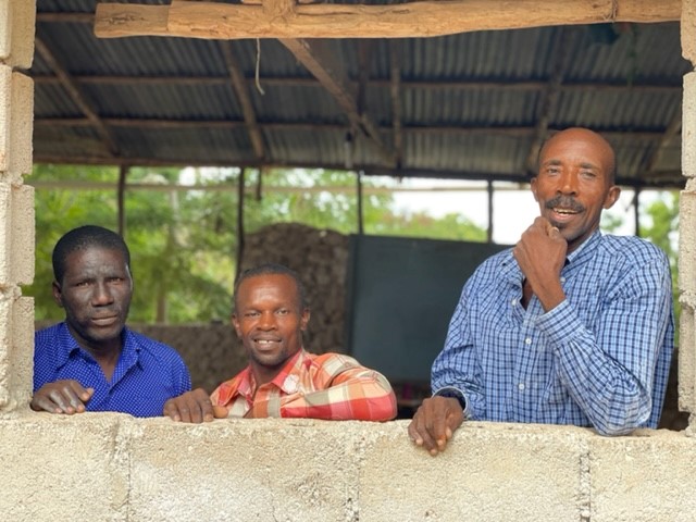 three men looking over a brick wall into the camera, smiling. 