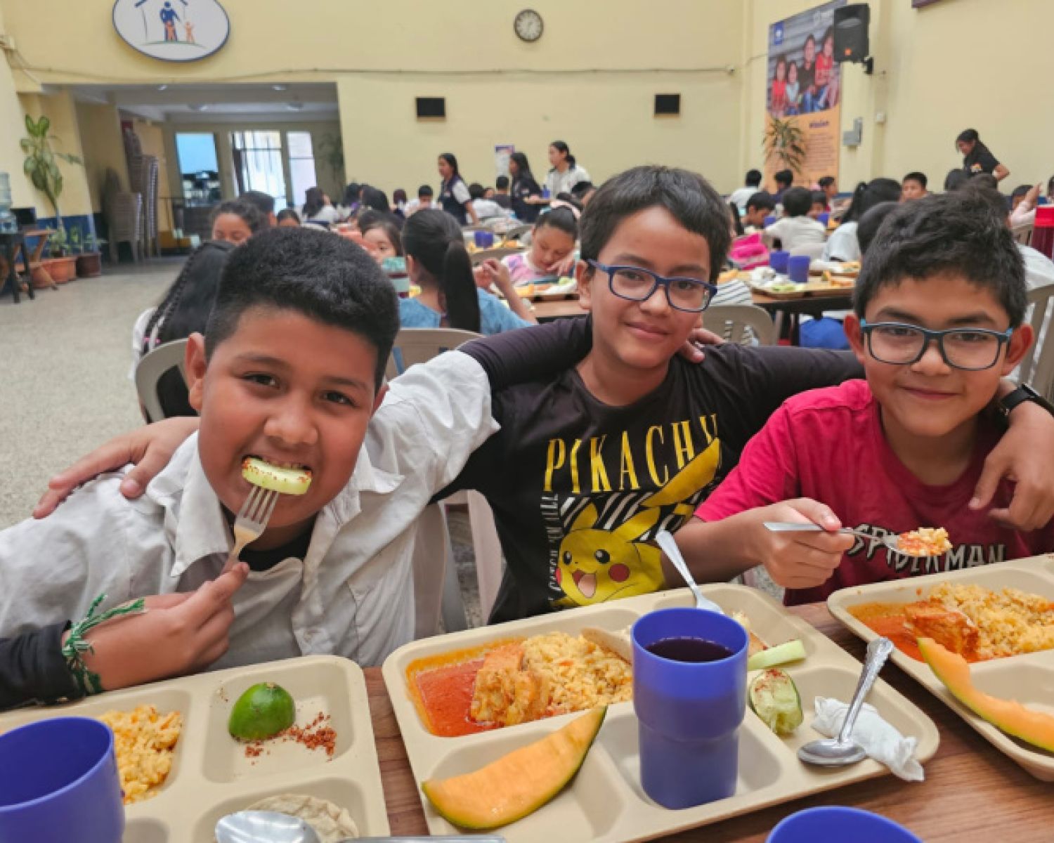 A group of young boys smiling for the camera whilst eating lunch at school 