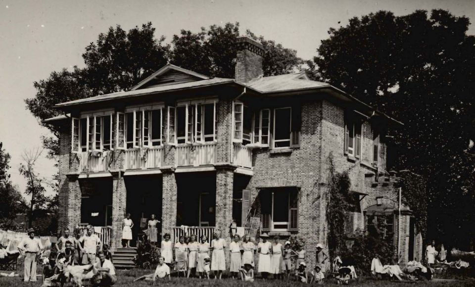 A vintage photograph of a large brick house with many people, including children, gathered in front of it, set in a lush, tree-filled landscape.
