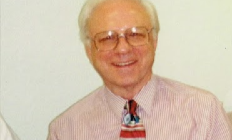 An elderly man with white hair and glasses is smiling at the camera. He is wearing a light pink, striped dress shirt and a colourful, patterned tie. The background is plain and light-coloured.