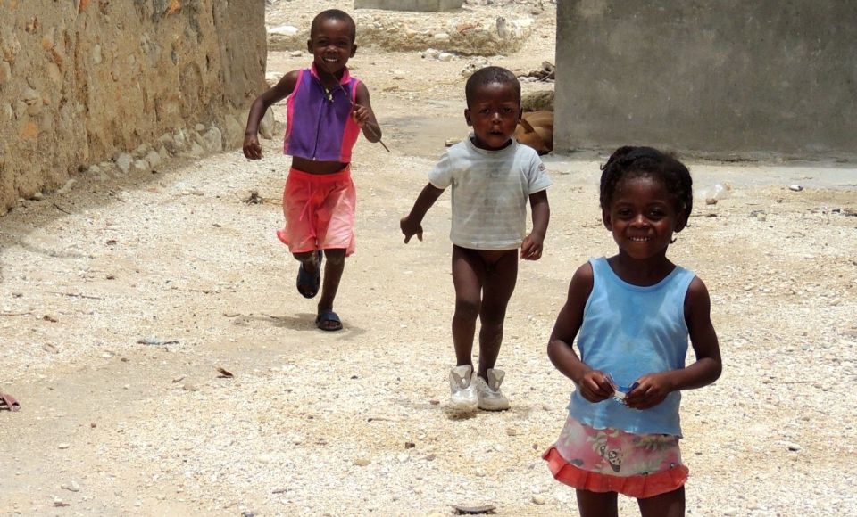 Three young children are playing and running on a dirt path between stone and concrete buildings. 