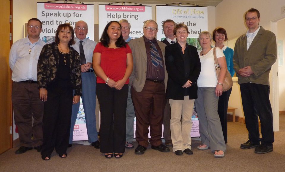 A group of ten adults is standing together indoors, posing for a photo in front of banners from worldshare.org.uk. 