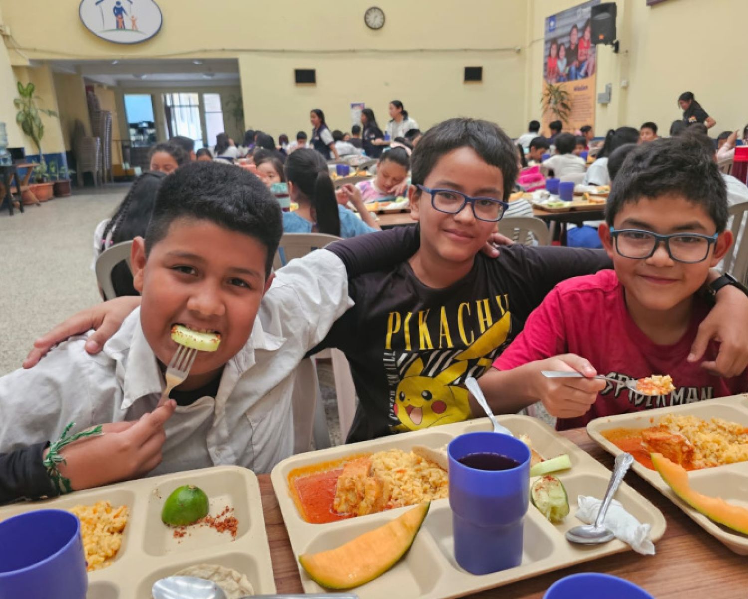 A group of young boys smiling for the camera whilst eating lunch at school 