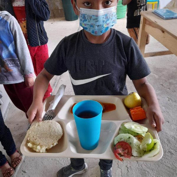 A young boy, with a lunch tray full of food, and a face mask on, looking into the camera.