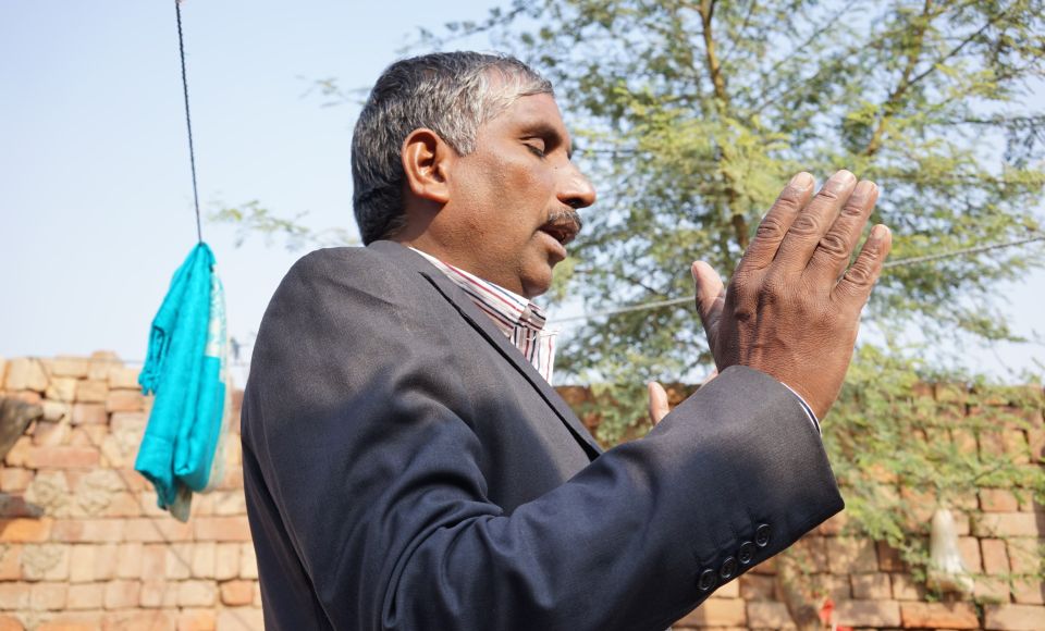 A man in a suit gestures with his hands while speaking outdoors, with a brick wall and tree in the background.