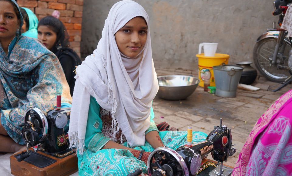 A young girl in a white headscarf and turquoise outfit sits at a sewing machine, with other women around her. 