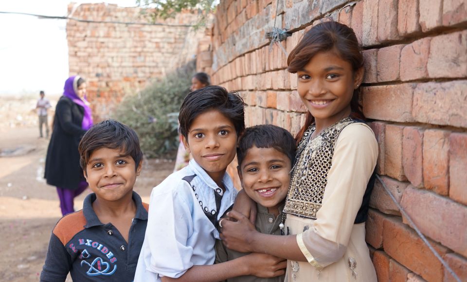 Four smiling children stand close together against a brick wall, with a few people and more bricks in the background.