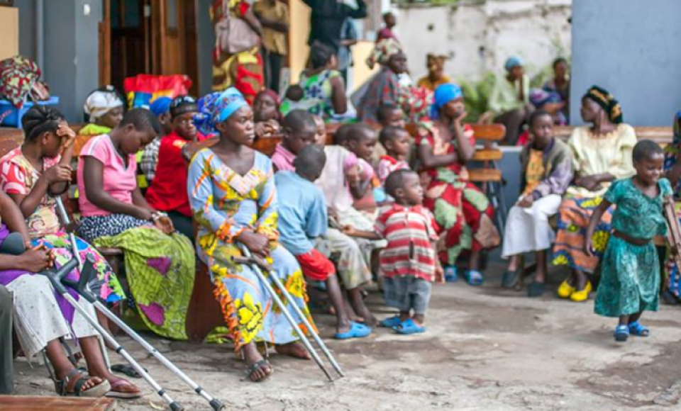 a large group of people sitting outside, a mixture of children and women. 