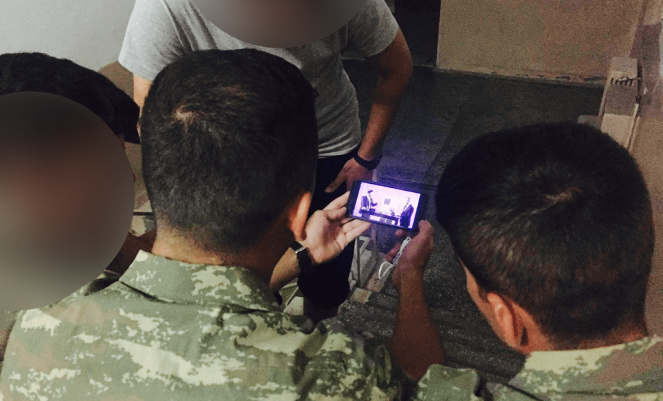 A group of army individuals huddled around a phone watching an interview. 