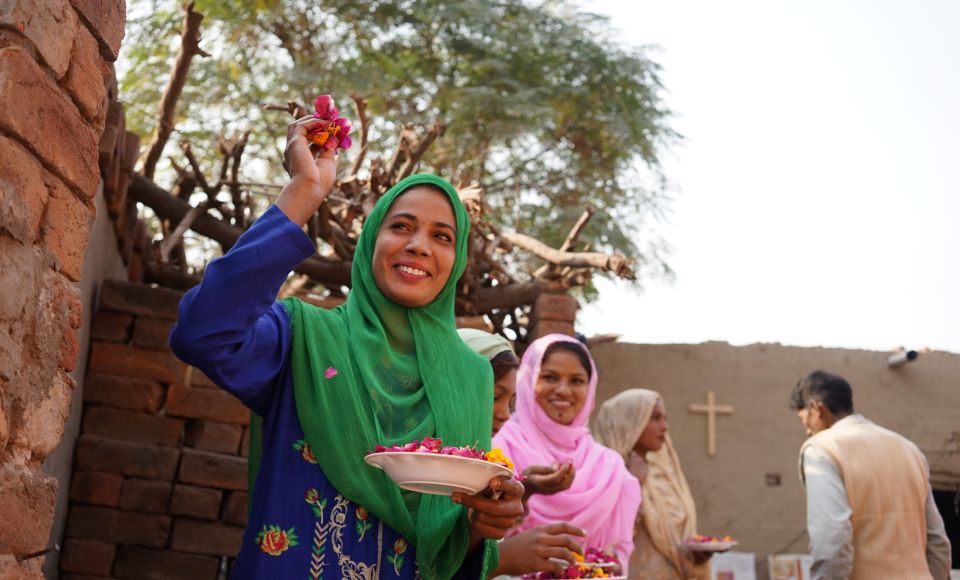 A smiling woman in a green headscarf holds a plate of flowers, with others around her, in an outdoor setting near a brick wall.