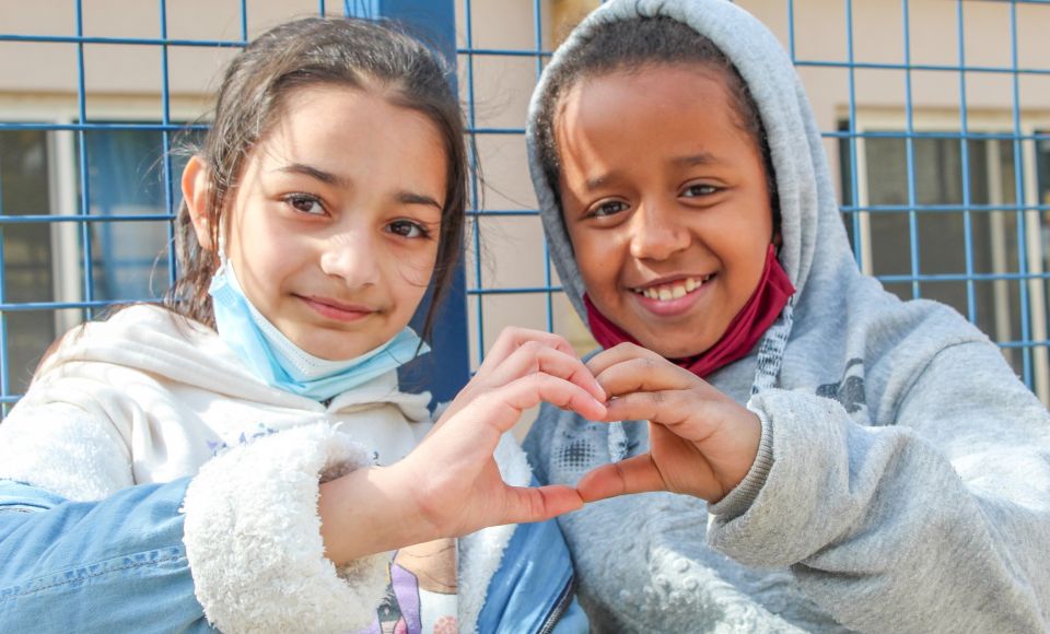 Two young girls smile and form a heart shape with their hands, wearing casual jackets and standing in front of a blue metal fence.