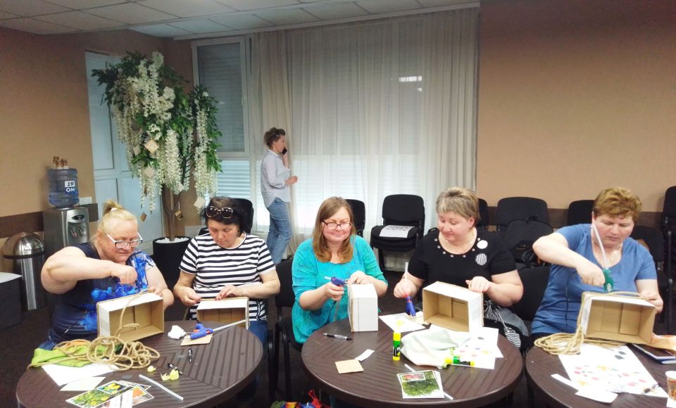 a group of older women practicing arts and crafts around a table. 