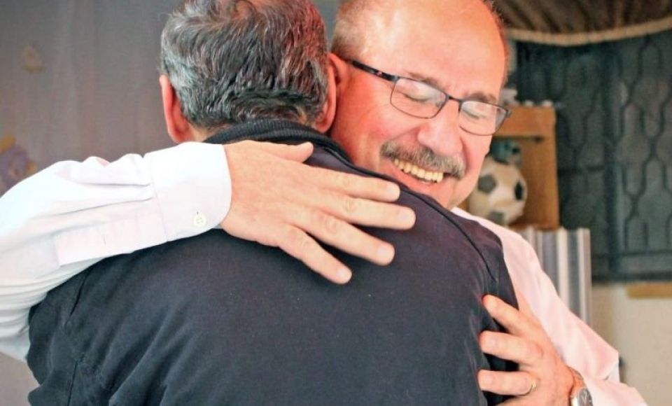 Two men warmly embrace, smiling with their eyes closed, in a heartfelt moment indoors.