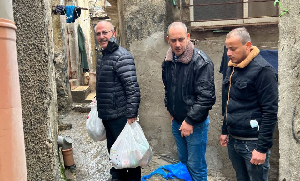 Three men stand in a narrow alleyway holding bags of supplies, wearing jackets on a chilly day. 