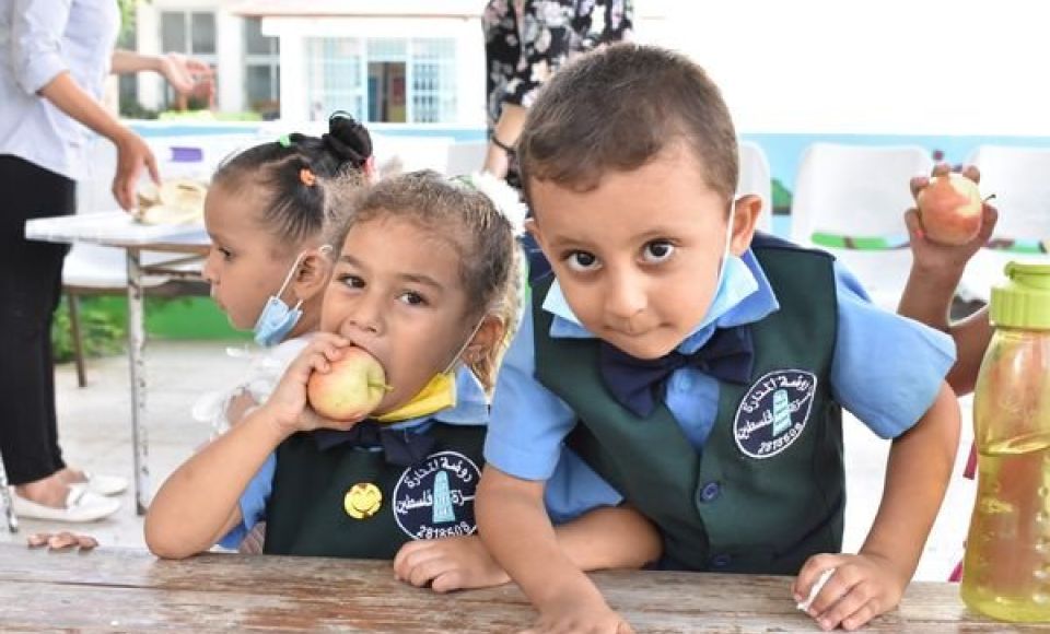 Two young children in school uniforms sit at a table, with one girl eating an apple and a boy looking curiously at the camera